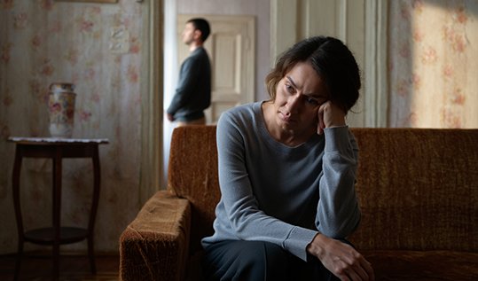Женщина сидит на диване дома, держась за голову, на заднем плане виден силуэт мужчины